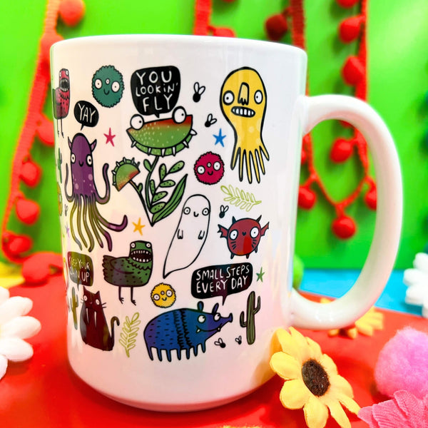 A cheerful white mug from Katie Abey, featuring a vibrant design of quirky monsters and encouraging words like "You lookin' fly" and "Small steps every day," placed against a lively green backdrop with red pom-poms and colourful daisies.