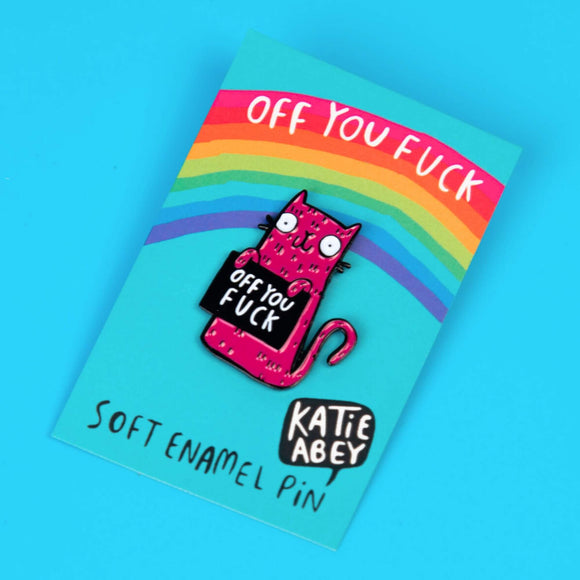 Katie Abey soft enamel pin featuring a quirky pink cat holding a sign that says 'Off You Fuck'. The pin is displayed on a blue card with a rainbow design and the text 'Off You Fuck' written at the top. The card also has the artist's name, Katie Abey, at the bottom.