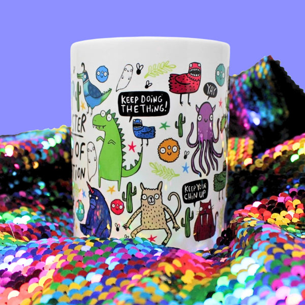 A fun and colourful Katie Abey mug featuring quirky, illustrated monsters and motivational messages like "Keep doing the thing!" and "Keep your chin up," placed on a dazzling, rainbow sequin background.