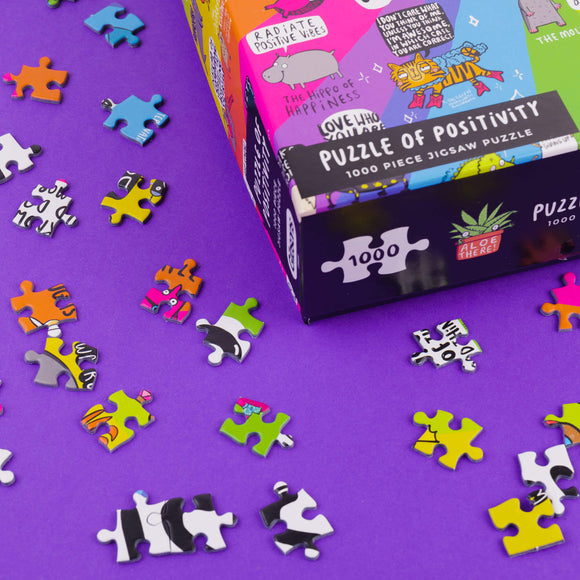 An image of a box with The Puzzle of positivity written in the middle with lots of fun illustrated characters on it by Katie Abey on a purple background there are also some loose puzzle pieces next to the box.