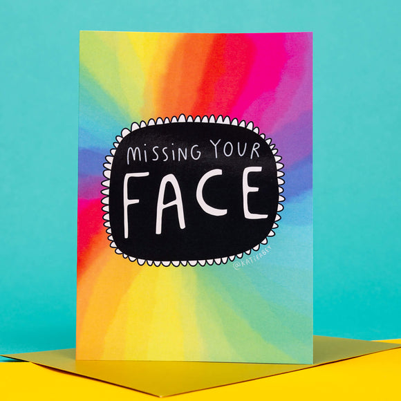 quirky rainbow coloured A6 greeting card with black circle in the centre with white writing on it that says 'missing your face'. The card comes with a gold envelope. Designed by Katie Abey.