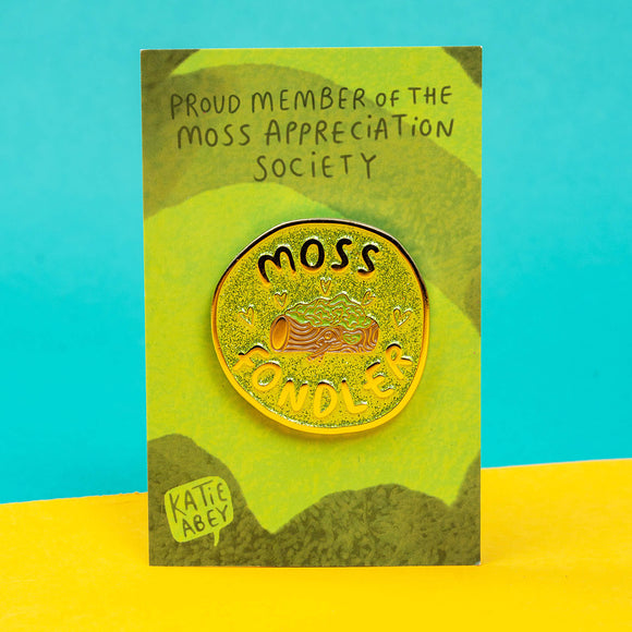 The Moss Fondler Enamel Pin Badge on its green gradient stripe backing card with top text reading 'proud member of the moss appreciation society' and Katie Abey's logo in the bottom left corner, its stood on a blue and yellow background. The glittery green circle enamel pin has a shiny gold coloured outline with a brown log in the centre covered in moss with hearts and text reading 'moss fondler' surrounding. Hand drawn design by Katie Abey for nature lovers.
