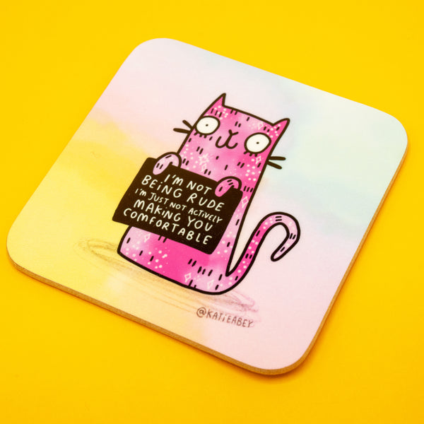 Angled product photo of the Katie Abey “I’m Not Being Rude” coaster featuring a pink illustrated cat holding a cheeky sign. The 9cm square coaster has rounded edges, a pastel background, and sits on a vibrant yellow surface. A fun and durable British-made coaster perfect for adding personality to your home or office.