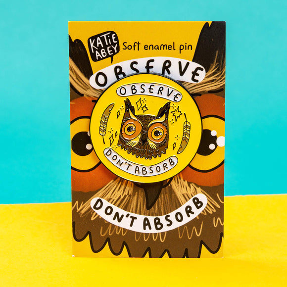 The Observe Don't Absorb Enamel Pin Badge on a brown owl backing card stood on a yellow and teal card background. The yellow base circle soft enamel pin has a brown smiley owl with big eyes surrounded by sparkles and two feathers with text in white banners reading 'observe don't absorb'.
