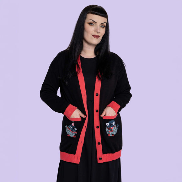 Front view of model wearing Katie Abey x Rebel Romance Pigeon Party Long Cardigan. Black longline cardigan with red trim, button front and pigeon illustrations on both pockets. Alternative UK fashion knit layered over black dress.
