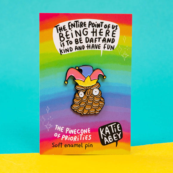 Katie Abey soft enamel pin featuring the 'Pinecone of Priorities' wearing a colourful jester hat and crown. The pin is displayed on a bright backing card with a rainbow background and the motivational phrase 'The entire point of us being here is to be daft and kind and have fun'. The artist's name, Katie Abey, is also featured at the bottom.