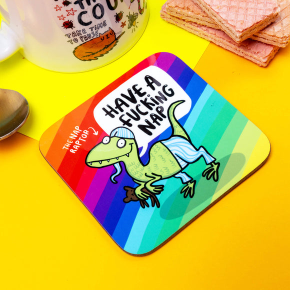 The coaster is a rounded corner square shape with a rainbow stripe background and a green raptor dinosaur smiling wearing pyjamas and holding a teddy bear. There's some text that read the 'The Nap Raptor' with an arrow pointing at the dinosaur and a speech bubble reading 'Have a fucking nap'.