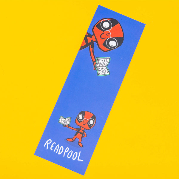 A blue bookmark with Deapool illustrated by Katie Abey on the front wearing glasses and reading a book. It says Readpool on the bottom