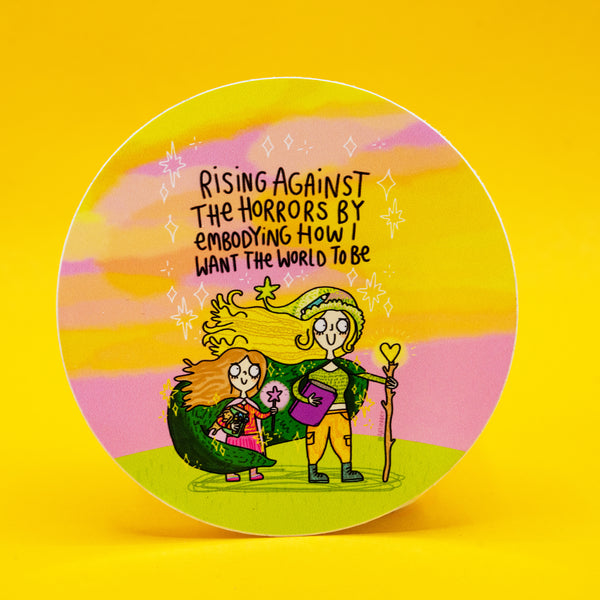 Circular pastel rainbow vinyl sticker by Katie Abey of a colourful illustrated adult and child both holding magical wands, standing together against a sunset sky. The sticker has a handwritten quote reading “Rising against the horrors by embodying how I want the world to be”. A joyful, empowering design encouraging kindness, creativity and being unapologetically yourself.