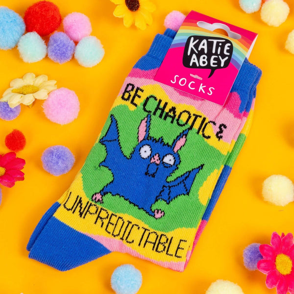 A pair of socks. The socks have wavy multi coloured chunks and a drawing by Katie Abey of a bat with text saying be chaotic & Unpredictable