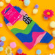 Back of the Be Chaotic & Unpredictable Rainbow Bat Socks laid on a yellow background amongst coloured flower and pom pom decorations. The socks have a wavy rainbow pattern.