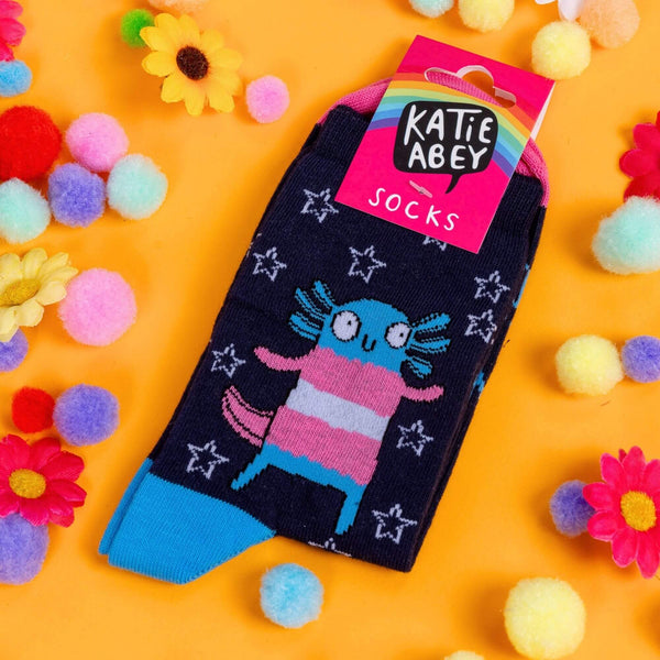 socks by Katie Abey with Axolotls in trans pride colours and stars with text saying don't tell me who I am on the front. The socks have pink toes and blue heels