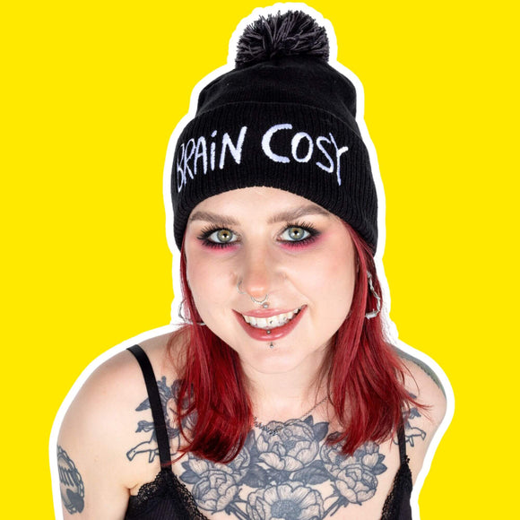 Brain cosy black knitted beanie bobble hat with grey pom pom and white embroidered lettering worn by a tattooed model with red hair and eye makeup against a bright yellow backdrop. Designed by Katie Abey in the UK