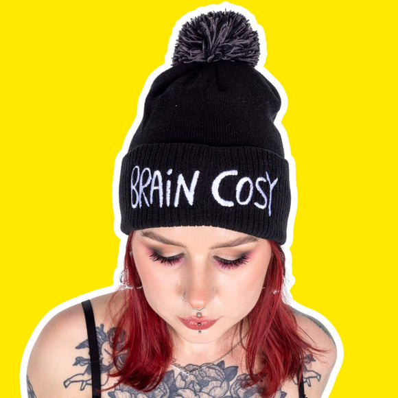 Brain cosy black knitted beanie bobble hat with grey pom pom and white embroidered lettering worn by a tattooed model with red hair and eye makeup against a bright yellow backdrop. Designed by Katie Abey in the UK