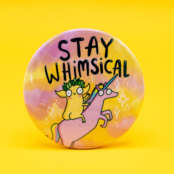 Round 58mm pin-back badge featuring an illustration by Katie Abey. The design shows a whimsical creature with green leafy hair riding a pink unicorn with a rainbow horn, set against a dreamy pastel background. Bold black lettering reads “STAY WHIMSICAL.” A fun, quirky accessory by Katie Abey, a small UK business encouraging you to embrace your weirdness.