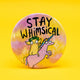 Round 58mm pin-back badge featuring an illustration by Katie Abey. The design shows a whimsical creature with green leafy hair riding a pink unicorn with a rainbow horn, set against a dreamy pastel background. Bold black lettering reads “STAY WHIMSICAL.” A fun, quirky accessory by Katie Abey, a small UK business encouraging you to embrace your weirdness.