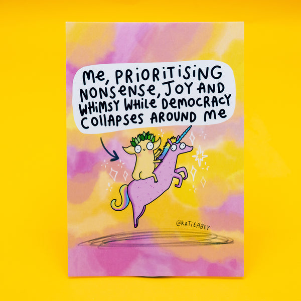 A6 postcard featuring a whimsical creature with leafy green hair riding a pink unicorn with a rainbow mane. Above, the hand-lettered text reads: “Me, prioritising nonsense, joy and whimsy while democracy collapses around me.” Set against a dreamy pastel background in yellow, pink, and purple. Designed by Katie Abey, this postcard is full of playful defiance and magical chaos.