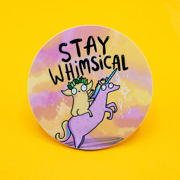 Round vinyl sticker featuring a quirky creature riding a pink unicorn with a rainbow mane, set against a dreamy pastel sky. The words “Stay Whimsical” are written above in bold black text. Approx. 8cm in diameter, this colourful, high-quality sticker is by Katie Abey, a small UK illustrator encouraging you to embrace your weirdness with fun and positivity.