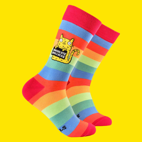  A close-up of Katie Abey's quirky "Absolute Bollocks" socks, featuring bold rainbow stripes and a yellow cat holding a sign that reads "Absolute Bollocks". These vibrant socks are perfect for adding a pop of colour and humour to your outfit, celebrating weirdness and individuality.