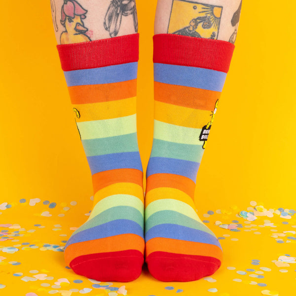 Front view of feet wearing the socks, displaying vibrant rainbow colours and a whimsical, cheeky cat design on both socks, with confetti spread on the yellow floor.