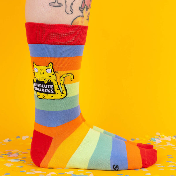 Colourful rainbow socks featuring a sweary yellow cat holding a sign that reads “Absolute Bollocks,” styled on tattooed legs, surrounded by bright yellow backdrop and confetti for a fun, quirky vibe.