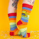 Colourful rainbow socks featuring a sweary yellow cat holding a sign that reads “Absolute Bollocks,” styled on tattooed legs, surrounded by bright yellow backdrop and confetti for a fun, quirky vibe.