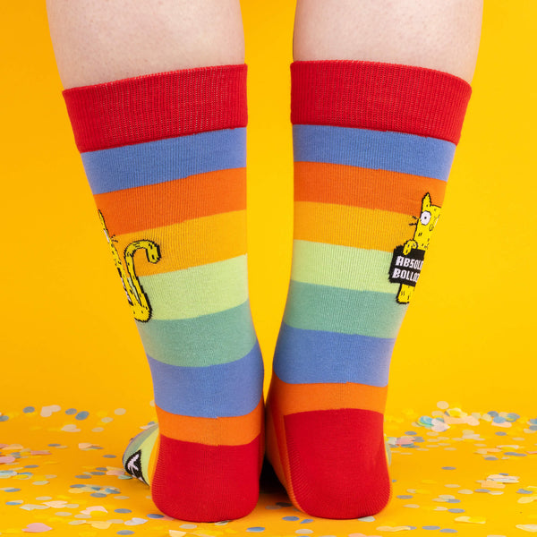 Back view of rainbow-striped socks, showing the colourful bands and a peek of the sock label, “Katie Abey Socctopus,” with confetti surrounding the feet.