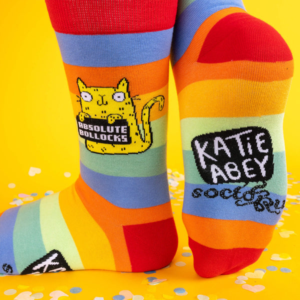 Close-up of brightly coloured rainbow socks featuring a yellow cat holding a sign that says “Absolute Bollocks,” worn on tattooed legs with a vibrant yellow background and confetti scattered on the floor.