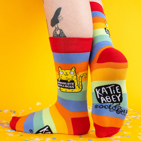 A fun, angled shot of the rainbow socks in action, with the yellow “Absolute Bollocks” cat front and centre, surrounded by a cheerful yellow backdrop and party-ready confetti.