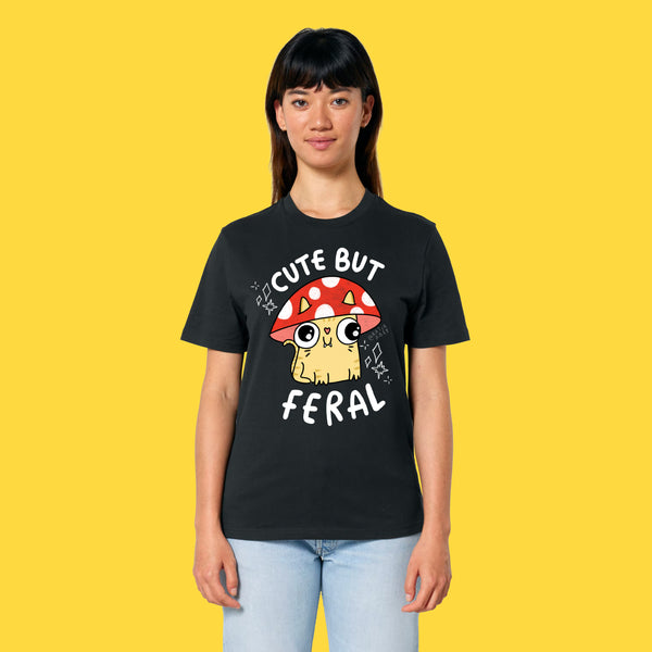 Model wearing the black Cute But Feral Cat adults t shirt. Front view showing cute illustrated cat artwork with mushroom hat and playful slogan. Unisex organic cotton t shirt styled casually against a yellow background.