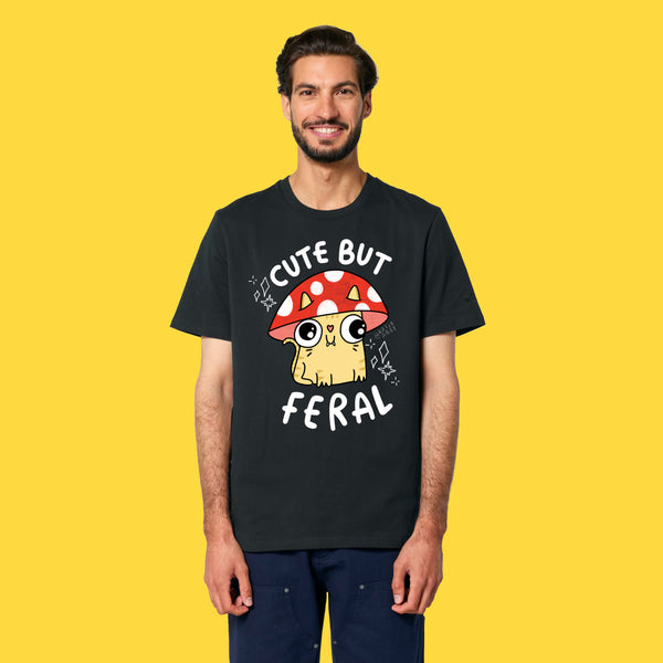 Model wearing the black Cute But Feral Cat adults t shirt by Katie Abey. Front view showing bold cat illustration and Cute But Feral slogan. Unisex graphic tee made from soft organic cotton, photographed on a yellow background.
