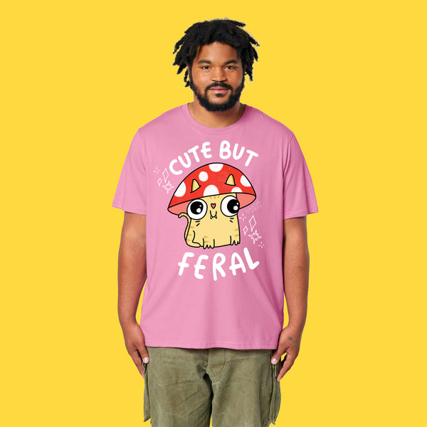 Model wearing the bubble pink Cute But Feral Cat adults t shirt. Front view showing playful Katie Abey cat artwork with mushroom hat and Cute But Feral slogan. Relaxed unisex fit organic cotton t shirt styled with green trousers on a yellow background.
