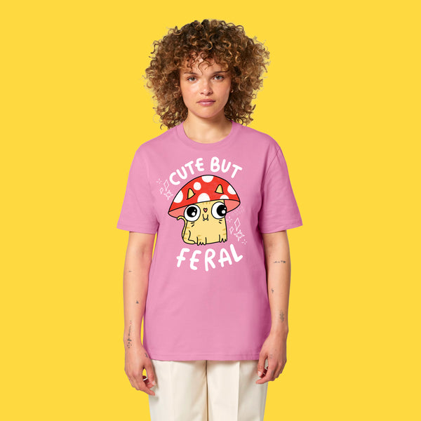 Model wearing the bubble pink Cute But Feral Cat adults t shirt by Katie Abey. Front view showing the cute illustrated cat in a red spotted mushroom hat with bold white Cute But Feral text. Unisex organic cotton tee photographed against a yellow background.