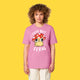 Model wearing the bubble pink Cute But Feral Cat adults t shirt by Katie Abey. Front view showing the cute illustrated cat in a red spotted mushroom hat with bold white Cute But Feral text. Unisex organic cotton tee photographed against a yellow background.