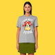 Model wearing the heather grey Cute But Feral Cat adults t shirt by Katie Abey. Front view showing the cute illustrated cat in a mushroom hat with Cute But Feral text. Styled with a green skirt on a bright yellow background.