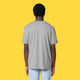 Model wearing the heather grey Cute But Feral Cat adults t shirt shown from the back. Loose fitting organic cotton tee with a casual drape, styled with light blue jeans on a yellow background.