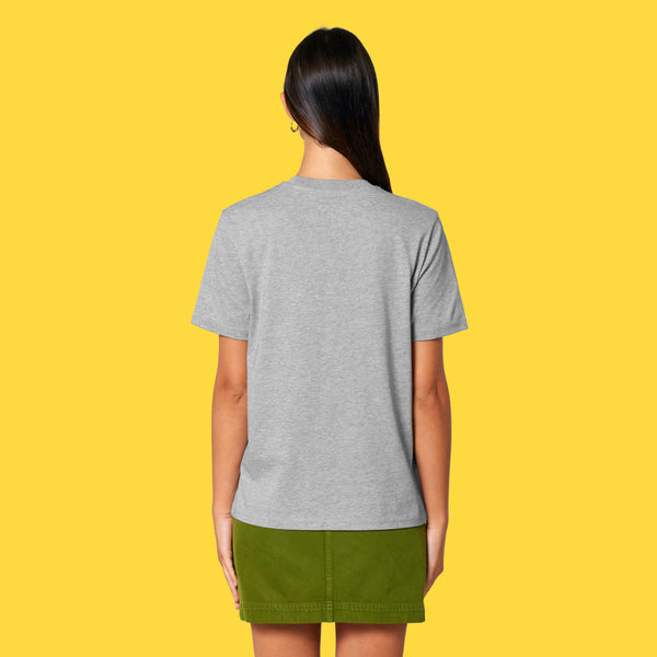 Model wearing the heather grey Cute But Feral Cat adults t shirt shown from the back. Relaxed unisex fit with short sleeves and crew neck, styled with a green skirt against a yellow background.