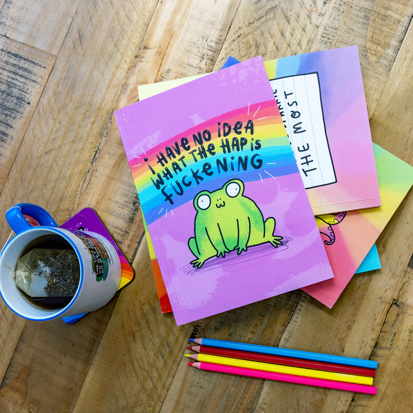 Katie Abey notebooks piled on top of each other on a wooden table next to a mug of tea and coloured pencils.