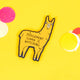 The Judgement Llama Magnet being held up over a yellow background. The yellow llama shaped magnet has a scowled expression with black text across its body reading 'judgement llama is watching'.