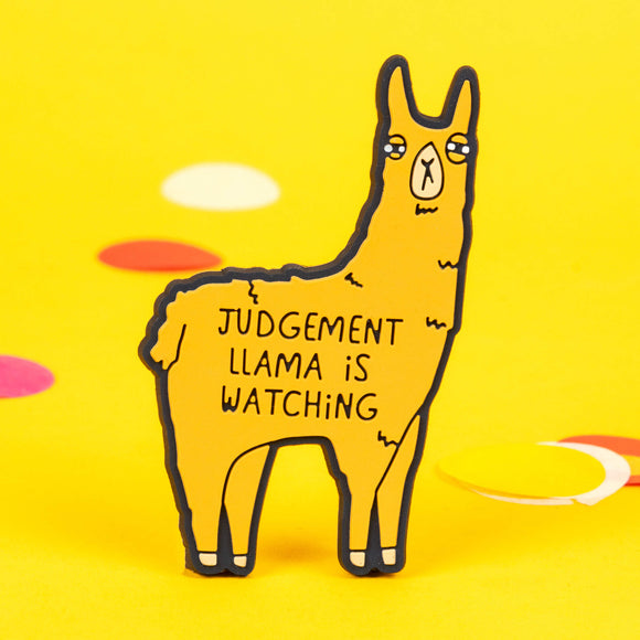 The Judgement Llama Magnet being held up over a yellow background. The yellow llama shaped magnet has a scowled expression with black text across its body reading 'judgement llama is watching'.