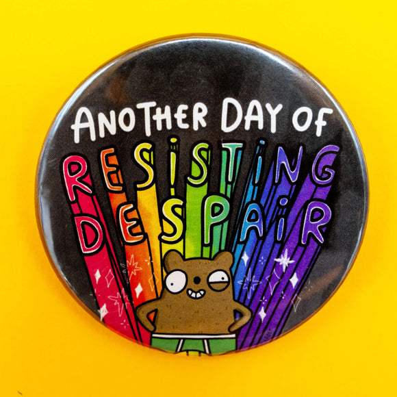 A round badge on a bright yellow background featuring the phrase "Another Day of Resisting Despair" in bold white and rainbow lettering. A brown cartoon bear character stands proudly at the bottom, with rainbow rays and sparkles behind them.