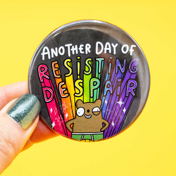 Close-up of a hand holding a round Katie Abey badge with the words "Another Day of Resisting Despair" in rainbow colours. The badge features a cartoon bear with hands on hips and a determined expression, set against a black background with colourful rays and sparkles.