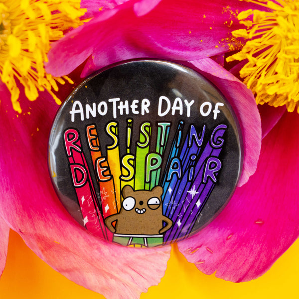 Katie Abey badge nestled in bright pink and yellow flower petals. The badge reads "Another Day of Resisting Despair" in rainbow text and shows a brown cartoon bear standing confidently with rainbow beams and stars behind them.