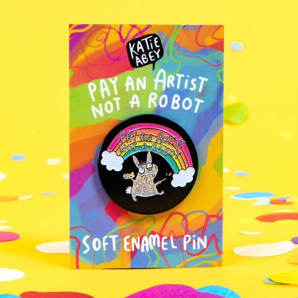 Pay an artist pin designed by Katie Abey in the UK. A black circle soft enamel pin with a sparkly grey rabbit wearing an artist hat and holding paint and brushes is smiling looking up at a rainbow reading 'pay an artist not a robot'. The pin is on a rainbow backing card with Katie Abey's logo at the top, then text that reads 'pay an artist not a robot' and bottom text that reads 'soft enamel pin'. 