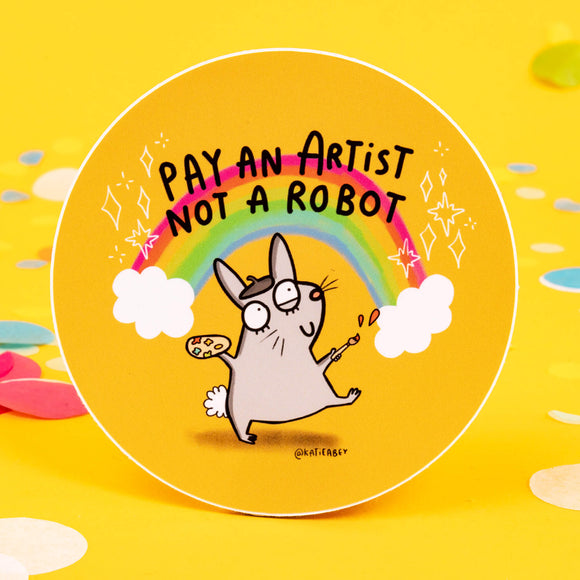 A sticker on a yellow background with a drawing by Katie Abey of a rabbit with a paint brush and a rainbow behind it with text saying pay an artist not a robot