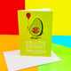 Here is your birthday avocardo A6 greeting card designed and printed in the UK by Katie Abey. The green base front cover features an avocado with arms, legs and a smiling face holding a balloon. The underneath text reads 'here is your birthday avocardo' with the card being underlined as the pun. The card is stood up 