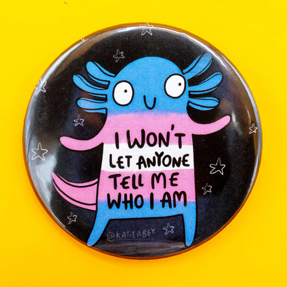 Round badge on a yellow background featuring a cartoon axolotl in the colours of the trans flag. Surrounded by small stars, the text on its body reads: “I won’t let anyone tell me who I am.”