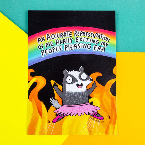 A postcard of a badger in a pink ballet outfit giving two fingers up on a black and fiery illustration with a rainbow above them saying An accurate representation of me finally exiting my people please era. Illustrated by Katie Abey