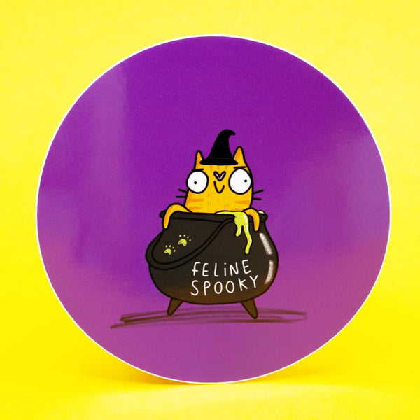Illustration of a cute orange cat wearing a witch hat, popping out of a bubbling black cauldron with green goo and the words Feline Spooky on the front. The vinyl sticker has a purple circular background and is created by Katie Abey, a UK illustrator known for quirky, weird, uplifting designs. Perfect for decorating laptops, notebooks, and gifts.