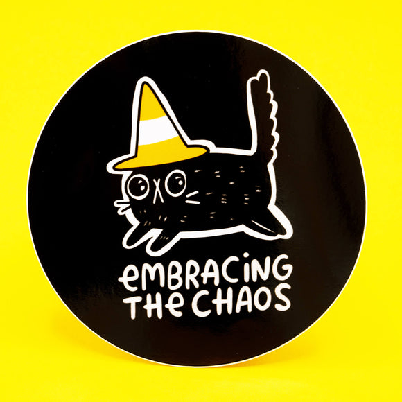 A round black vinyl sticker by Katie Abey featuring a quirky black cat wearing a yellow and white traffic cone hat with the words “Embracing The Chaos” below. Printed in bold white line art with a playful, weird vibe. Premium durable vinyl, 10cm size, perfect for laptops, notebooks, and water bottles. Bright yellow background.